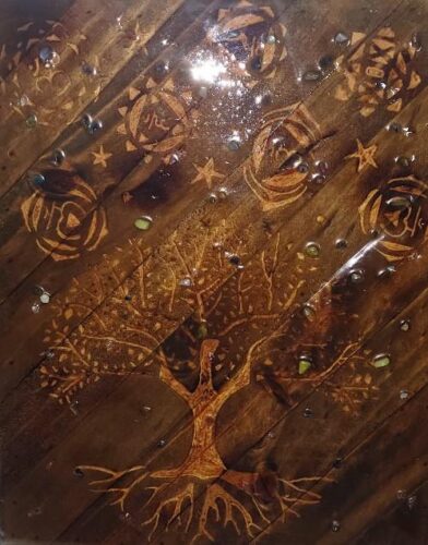 Overhead view of a wooden surface made of diagonal planks, etched and burned with symbolic designs. A large tree with spreading branches and visible roots is carved into the lower section, while stars, circular motifs, and abstract symbols appear above. Small glass pebbles are scattered across the surface, and light reflects near the top center.