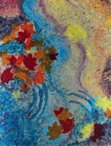 A textured multimedia or collage painting depicting colorful, star-shaped leaves floating down a moving stream. The artwork is created on a coarse, fibrous paper surface that gives the entire piece a speckled, granular quality. On the left side, a swirling blue field representing water flows downward, carrying a dense cluster of autumn-colored leaves in shades of crimson red, orange, and golden brown. A few stray red and brown leaves drift into the lower right corner. The right side of the composition transitions into a warm, sandy field of mottled gold, beige, and light yellow, separated from the blue water by faint, wavy strokes of purple and bright yellow paint.