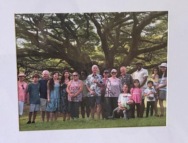 A wide group photograph of approximately fifteen people of diverse ages and ethnicities posing together outdoors on a green lawn. They are standing in a long line in front of a massive, sprawling banyan or monkeypod tree with thick, twisting branches that create a dense canopy overhead. The group is dressed in casual, warm-weather attire, including t-shirts, summer dresses, and patterned button-downs. On the far right, a few young children stand with the adults, and near the center-right, a woman kneels on the grass while playfully holding a young child upside down. The lighting is soft and natural, and the photo is framed within a white mat board.